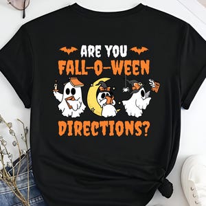 Puede incluir: Camiseta negra con un diseño de Halloween. El diseño presenta tres fantasmas de dibujos animados, una luna creciente y el texto "ARE YOU FALL-O-WEEN DIRECTIONS?" en naranja y blanco.
