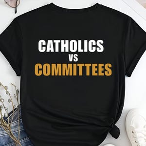 May include: Black t-shirt featuring the text "CATHOLICS VS COMMITTEES" in white and gold. The casual tee is likely made from cotton or a similar material. The design is centrally placed on the front of the garment.