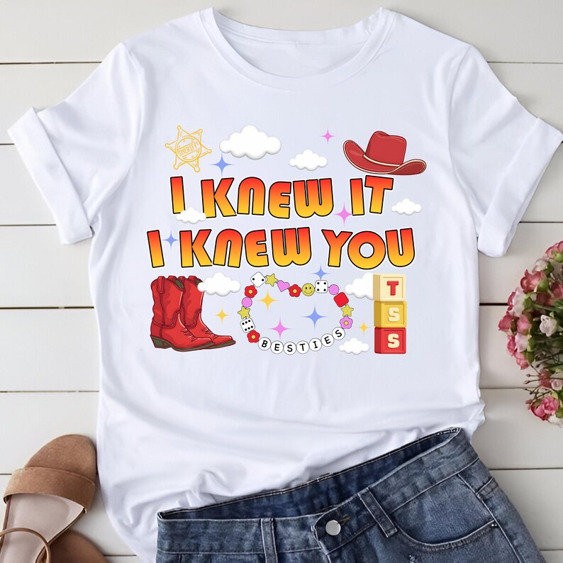 May include: A white t-shirt featuring a graphic print with the text I KNEW IT I KNEW YOU in yellow and orange. The design includes a red cowboy hat, red cowboy boots, a beaded bracelet with the word BESTIES, dice, flowers, and alphabet blocks.