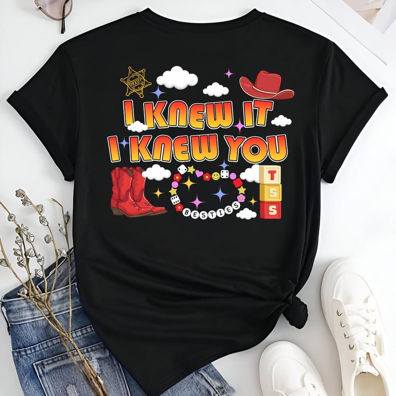 May include: A black t-shirt featuring a graphic print with the text I KNEW IT I KNEW YOU. The design includes a red cowboy hat, a sheriff star, red cowboy boots, a beaded necklace with the word BESTIES, dice, and three stacked blocks with TSS.