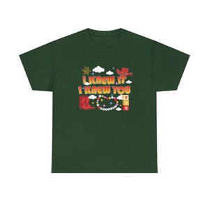May include: A dark green short-sleeve t-shirt featuring a graphic print with the text I KNEW IT I KNEW YOU. The design includes a red cowboy hat, a red cowboy boot, a sheriff star, white clouds, colorful beads, and stacked blocks with letters.