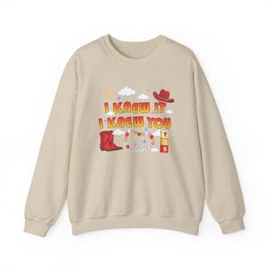 May include: A beige crewneck sweatshirt featuring a graphic print with the text I KNEW IT I KNEW YOU. The design includes a red cowboy boot, a red cowboy hat, a beaded necklace, three stacked blocks with the letters T S S, clouds, and stars.