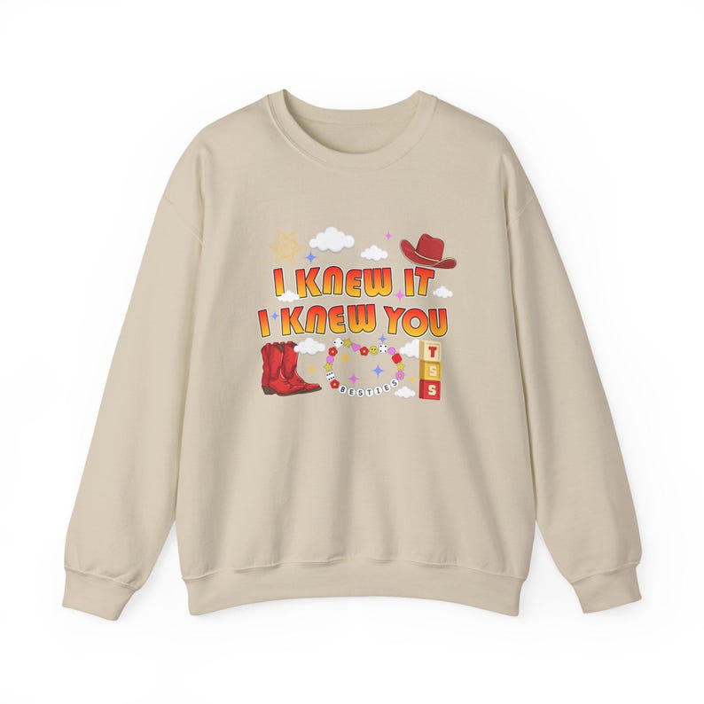 May include: A beige crewneck sweatshirt featuring a graphic print with the text I KNEW IT I KNEW YOU. The design includes a red cowboy boot, a red cowboy hat, a beaded necklace, three stacked blocks with the letters T S S, clouds, and stars.