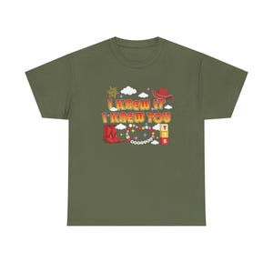 May include: An olive green t-shirt featuring a graphic print with the text I KNEW IT I KNEW YOU. The design includes a red cowboy hat, red cowboy boots, a sheriff star, white clouds, colorful stars, a beaded necklace, and three stacked blocks.
