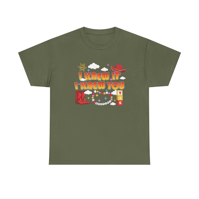 May include: An olive green t-shirt featuring a graphic print with the text I KNEW IT I KNEW YOU. The design includes a red cowboy hat, red cowboy boots, a sheriff star, white clouds, colorful stars, a beaded necklace, and three stacked blocks.