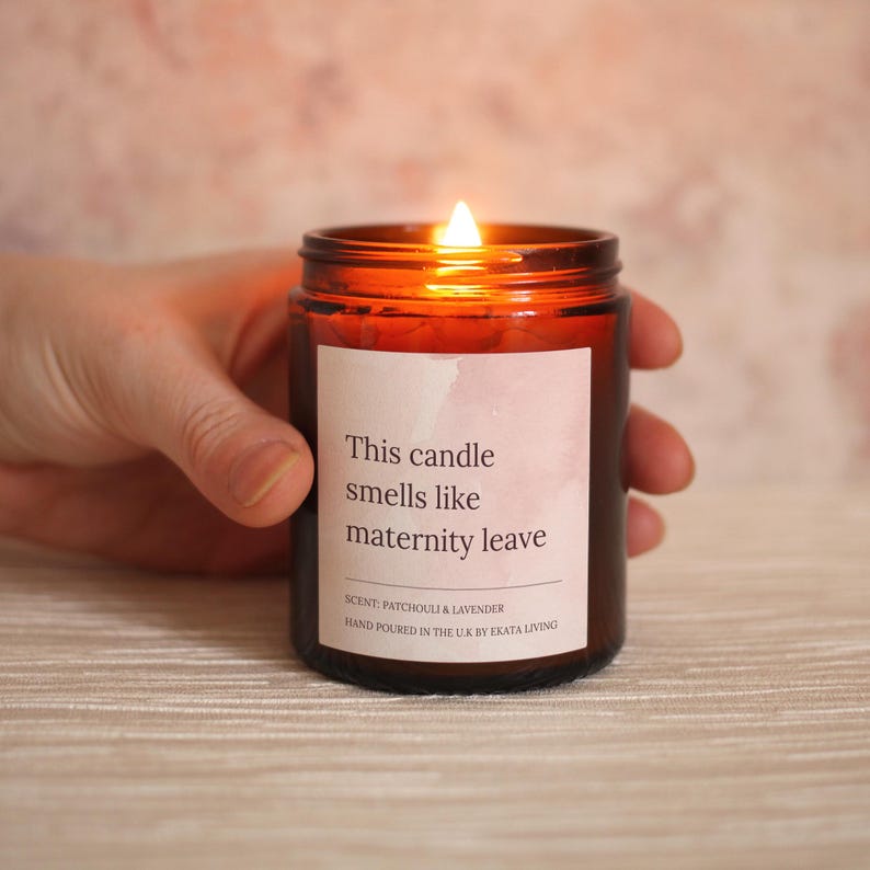 May include: A lit brown glass candle with a white label that reads "This candle smells like maternity leave." The scent is patchouli and lavender. The candle is hand-poured in the U.K. by Ekata Living.