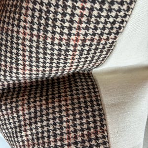 May include: Close-up of a pillow with a houndstooth pattern in brown, black, and red. The pillow has a cream-colored linen side. The fabric is textured and appears to be a woven material. The pillow is likely for home decor.