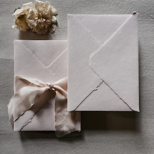 May include: Two off-white envelopes with a textured finish, one tied with a pale pink ribbon. A delicate, cream-colored flower rests near the envelopes. The envelopes have a unique, folded design, suggesting a handmade or artisanal quality.