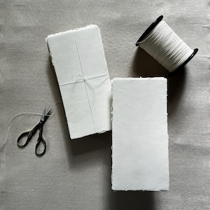 May include: A still-life image featuring stacks of white paper, bound with twine, alongside a spool of white thread and small black scissors. The paper has a textured, handmade appearance. The items are arranged on a gray surface.