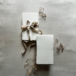 May include: Two stacks of handmade, deckle-edged paper cards. One stack is tied with a beige ribbon. Dried, delicate flowers are arranged around the cards. The background is a neutral, textured surface.