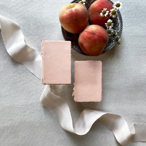 May include: A still-life arrangement featuring three ripe peaches in a decorative plate with small white flowers. Two pink rectangular cards and a cream-colored ribbon are also present, all set against a light gray background.