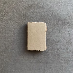 May include: A rectangular, off-white handmade paper block with rough, uneven edges. The paper has a natural, textured appearance, suggesting a craft or art supply. The block is centered against a grey, textured background.