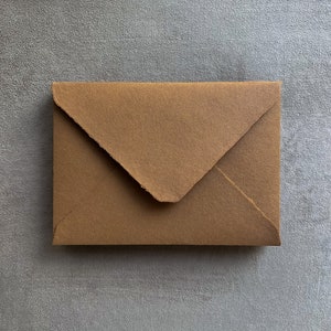 May include: A brown paper envelope with a textured surface. The envelope is rectangular with a triangular flap closure. The paper has a natural, organic look, suggesting it may be handmade or eco-friendly.