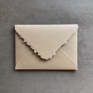 May include: A handmade, beige envelope with a torn edge. The envelope is rectangular and has a flap that is folded over. The paper has a textured appearance, and the background is a solid gray.