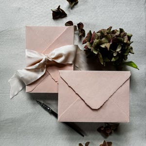 May include: Two blush pink envelopes with deckled edges, one tied with a cream-colored silk ribbon. A black fountain pen and dried flowers are arranged on a light gray surface, creating an elegant stationery theme.