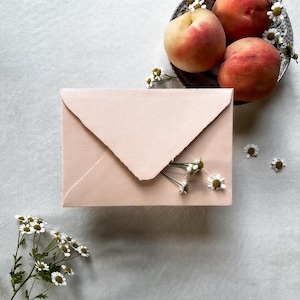 May include: A blush pink envelope with a torn edge, adorned with small white flowers, rests on a light gray surface. A decorative plate holds three ripe peaches, also decorated with white flowers. A still life composition.