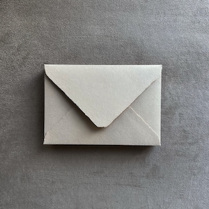 May include: A small, off-white envelope with a textured surface. The envelope is rectangular with a triangular flap. The paper has a slightly rough, handmade appearance. The envelope is centered on a gray background.