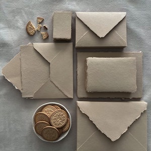 May include: A collection of beige stationery and biscuits is arranged on a grey surface. Several envelopes and notecards with rough edges are positioned around a small bowl filled with round biscuits. Some biscuits are broken.
