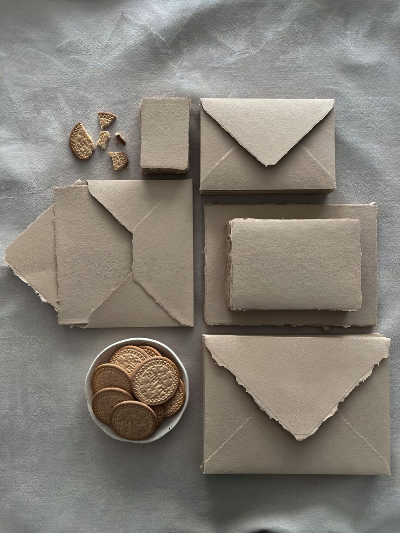 May include: A collection of beige stationery and biscuits is arranged on a grey surface. Several envelopes and notecards with rough edges are positioned around a small bowl filled with round biscuits. Some biscuits are broken.
