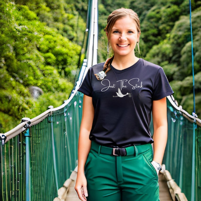 May include: A woman wearing a black t-shirt with the text "Just The Stork" and a white stork graphic. She is standing on a green suspension bridge.