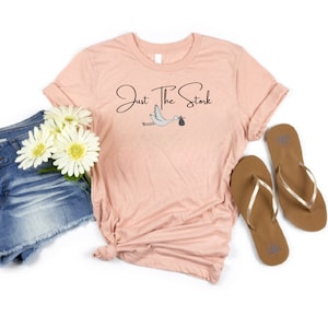May include: A light pink t-shirt with the text "Just The Stork" in black lettering and a cartoon stork carrying a baby in a bag. The shirt is laying on a white surface with a pair of brown flip flops and a pair of denim shorts with white flowers.