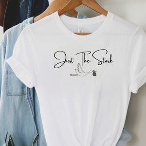 May include: White t-shirt with the text "Just The Stork" in black cursive font and a black and white illustration of a stork carrying a baby in a bag.
