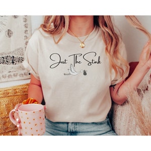 May include: A white t-shirt with the text "Just The Stork" in black cursive font. A black and white illustration of a stork carrying a baby in a bag is below the text.