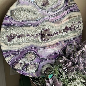 May include: Round resin art piece with a purple and white agate design. The resin is clear and the agate is embedded in the resin. The agate is a mix of purple and white crystals.