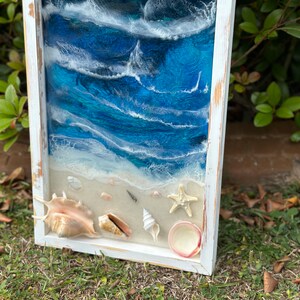 May include: A framed resin art piece depicting a beach scene with blue and white waves, a sandy beach, and seashells. The frame is white with a distressed finish.