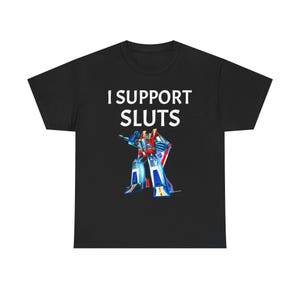 Peut inclure: T-shirt noir avec l'inscription blanche "I SUPPORT SLUTS". En dessous, une illustration colorée d'un robot rouge, bleu et blanc. Le robot est dans une pose dynamique.
