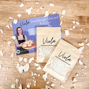 Puede incluir: Dos bolsas de papel marr&oacute;n con texto blanco que dice "Viola Dehydrated Sourdough Starter". Las bolsas est&aacute;n sobre una superficie de madera con trozos de papel blanco esparcidos a su alrededor. Una tarjeta de receta con el texto "Viola Sourdough Discard Biscuits" est&aacute; en el fondo.