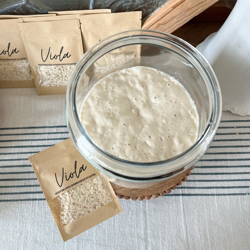 Puede incluir: Un tarro de vidrio lleno de masa madre blanca, sentado sobre una base de madera. Un peque&ntilde;o paquete de papel marr&oacute;n con el texto "Viola Masa Madre Deshidratada" es visible en primer plano.