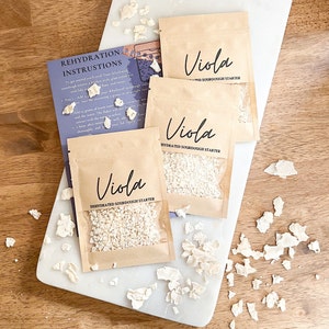 Puede incluir: Tres peque&ntilde;os paquetes de papel marr&oacute;n con ventanas de pl&aacute;stico transparente, cada uno contiene un iniciador de masa madre deshidratado blanco. Los paquetes est&aacute;n etiquetados como "Viola Iniciador de masa madre deshidratado" y "Viola Iniciador de masa madre hidratado". Los paquetes est&aacute;n sobre una superficie de madera marr&oacute;n claro con trozos de iniciador de masa madre deshidratado blanco esparcidos a su alrededor.