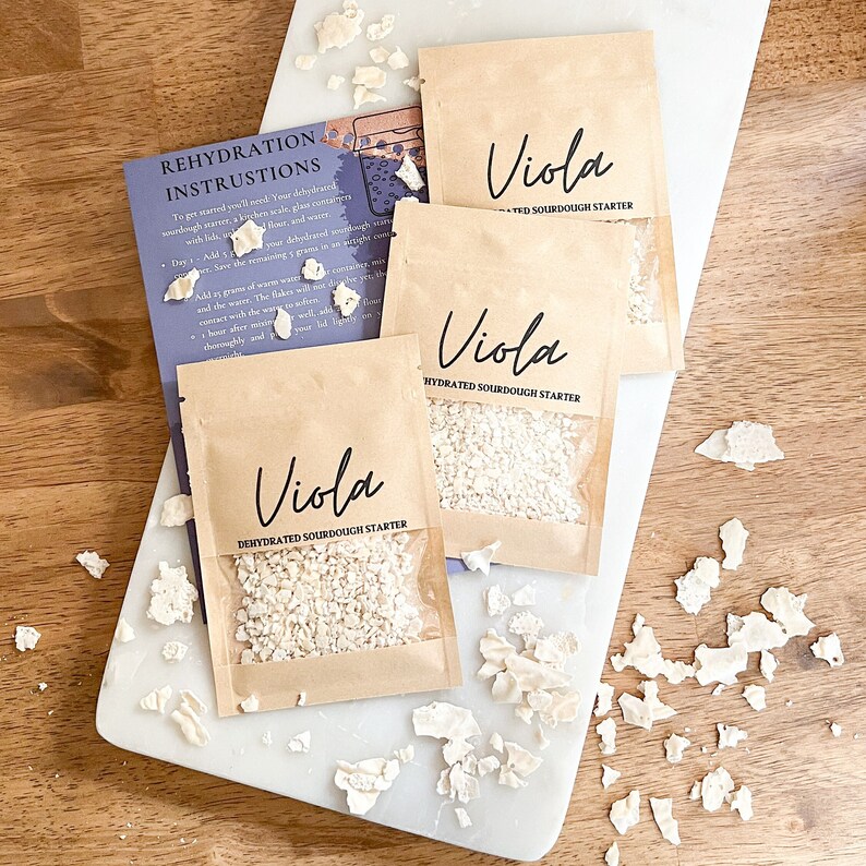 Puede incluir: Tres peque&ntilde;os paquetes de papel marr&oacute;n con ventanas de pl&aacute;stico transparente, cada uno contiene un iniciador de masa madre deshidratado blanco. Los paquetes est&aacute;n etiquetados como "Viola Iniciador de masa madre deshidratado" y "Viola Iniciador de masa madre hidratado". Los paquetes est&aacute;n sobre una superficie de madera marr&oacute;n claro con trozos de iniciador de masa madre deshidratado blanco esparcidos a su alrededor.