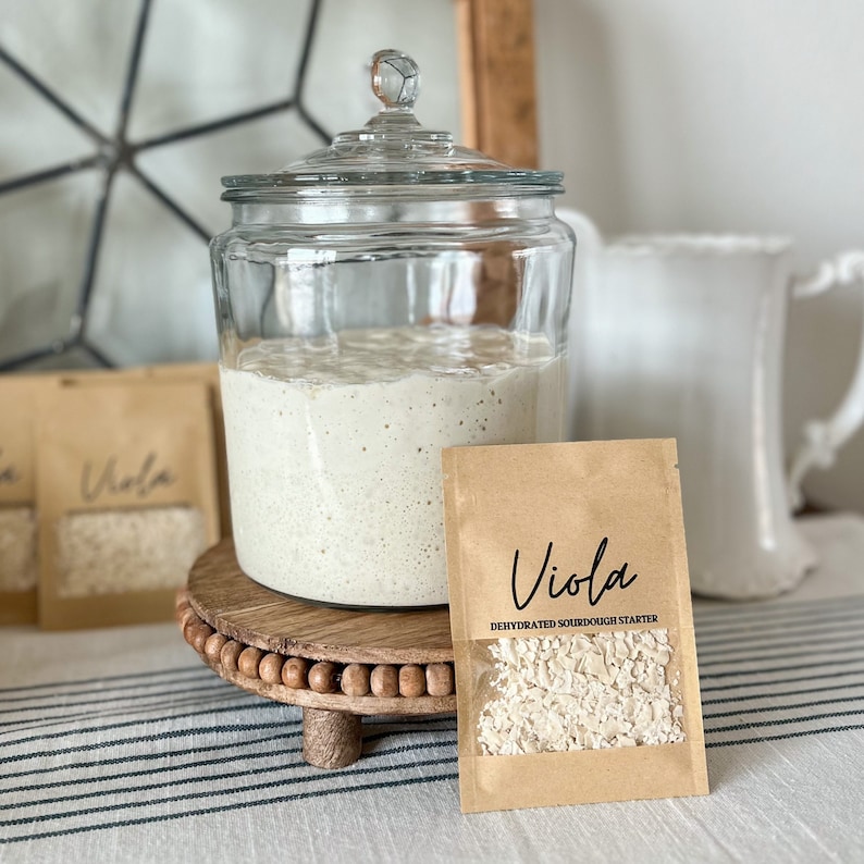 Puede incluir: Un tarro de vidrio con tapa lleno de masa madre blanca. Un paquete de papel marr&oacute;n con una ventana transparente muestra un iniciador de masa madre deshidratado con el texto "Viola Iniciador de masa madre deshidratado".