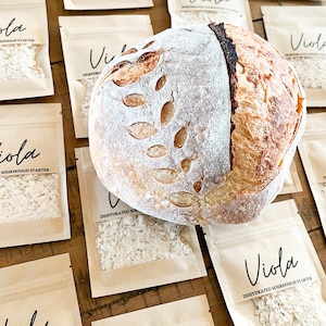 Puede incluir: Un pan de masa madre con un dise&ntilde;o de hojas en la parte superior, sentado en una mesa con varias bolsas peque&ntilde;as de papel marr&oacute;n que contienen masa madre deshidratada. Las bolsas est&aacute;n etiquetadas con "Viola Dehydrated Sourdough Starter".