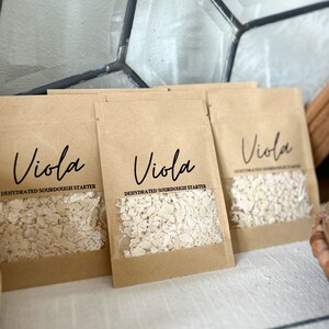 Puede incluir: Tres paquetes de papel marr&oacute;n con una ventana transparente que muestra masa madre deshidratada en el interior. Cada paquete tiene la palabra "Viola" impresa en el frente, junto con el texto "Masa madre deshidratada".