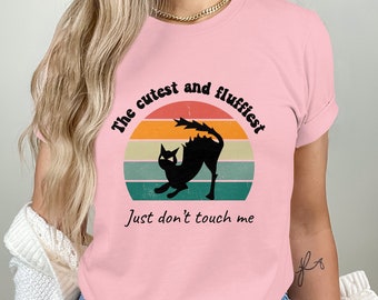 May include: Pink t-shirt with a black cat silhouette in a retro sunset design. The text reads "The cutest and fluffiest Just don't touch me".