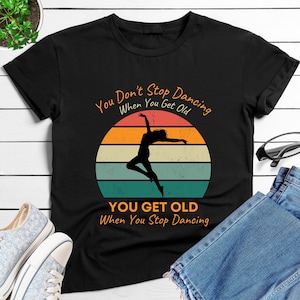 Peut inclure: T-shirt noir avec un motif rétro représentant une silhouette de danseuse devant un coucher de soleil. Le texte dit : "You Don't Stop Dancing When You Get Old, You Get Old When You Stop Dancing."