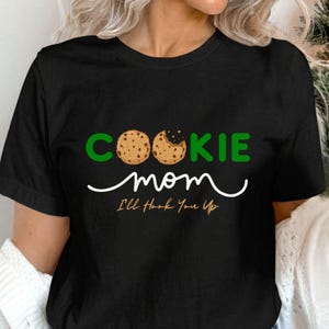 May include: Black t-shirt with the words "COOKIE mom" in green and white. The word "COOKIE" has two cookie images as the letters "O" and "O". Below the words "COOKIE mom" is the phrase "I'll Hook You Up".