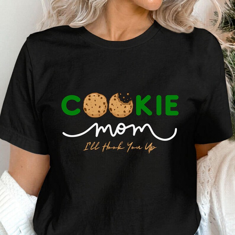 May include: Black t-shirt with the words "COOKIE mom" in green and white. The word "COOKIE" has two cookie images as the letters "O" and "O". Below the words "COOKIE mom" is the phrase "I'll Hook You Up".