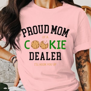 May include: A light pink t-shirt with the text "PROUD MOM of a COOKIE DEALER" in black and green. Two cookie illustrations are between the words "COOKIE" and "DEALER". The phrase "I'LL HOOK YOU UP" is below the main text.