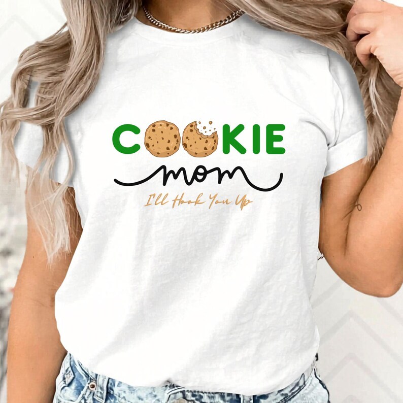 May include: White t-shirt with the words "COOKIE mom" in green and black, with a cookie graphic. Below the words is the phrase "I'll Hook You Up" in a handwritten style font. The shirt is a casual style.