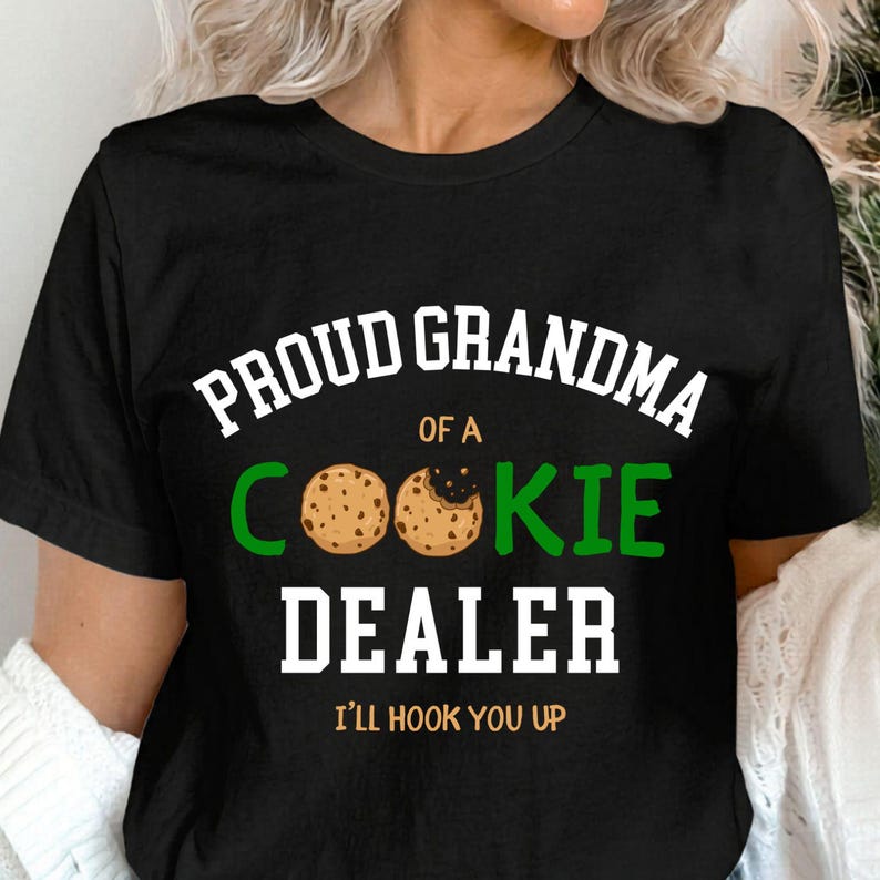May include: Black t-shirt with the text "PROUD GRANDMA OF A COOKIE DEALER" in white and green. The word "COOKIE" features two cookie graphics. The phrase "I'LL HOOK YOU UP" is printed at the bottom in gold.