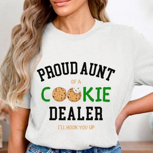 May include: White t-shirt with the text "PROUD AUNT of a COOKIE DEALER" in black and green. Two cookie illustrations are featured. The phrase "I'LL HOOK YOU UP" is printed below the main text.