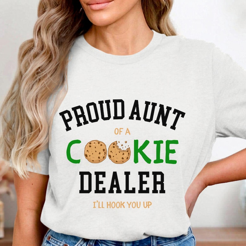 May include: White t-shirt with the text "PROUD AUNT of a COOKIE DEALER" in black and green. Two cookie illustrations are featured. The phrase "I'LL HOOK YOU UP" is printed below the main text.