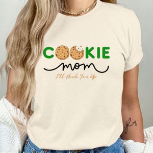 May include: Cream-colored t-shirt with the words "COOKIE mom" in green and black, featuring a cookie graphic. Below the text is the phrase "I'll Hook You Up" in orange.