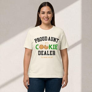 May include: A cream-colored t-shirt with the text "PROUD AUNT OF A COOKIE DEALER" in black and green. The word "COOKIE" features a cookie graphic. Below the text is "I'LL HOOK YOU UP" in black. The shirt is worn by a person.