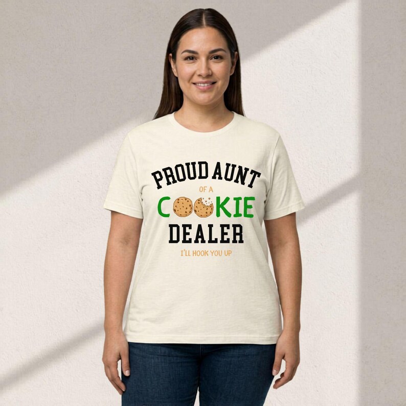May include: A cream-colored t-shirt with the text "PROUD AUNT OF A COOKIE DEALER" in black and green. The word "COOKIE" features a cookie graphic. Below the text is "I'LL HOOK YOU UP" in black. The shirt is worn by a person.