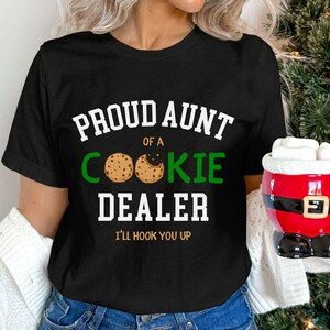 May include: Black t-shirt with the text "PROUD AUNT OF A COOKIE DEALER" in white and green. The design includes cookie graphics and the phrase "I'LL HOOK YOU UP" in white. A Santa mug is held in the background.